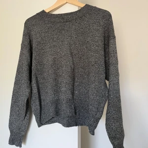 Glittrig grå stickad tröja - En glittrig grå stickad tröja med rund halsringning och ribbade muddar vid ärmslut och nederkant. Tröjan har en avslappnad passform och är perfekt för lager-på-lager-stil. Materialet känns mjukt och bekvämt.