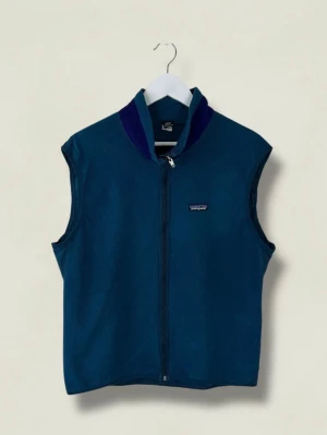 Blå fleeceväst från Patagonia - Mörkblå fleeceväst från Patagonia med hel dragkedja framtill, hög krage med marinblå detalj och liten logga på bröstet. Praktisk ficka med dragkedja på ryggen. Perfekt för lager-på-lager och outdoor-stil.