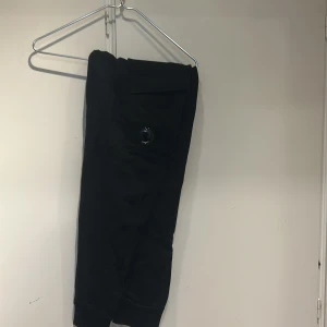 Svarta mjukisbyxor med ficka - Svarta mjukisbyxor med en stor ficka på ena benet och en dekorativ detalj i form av en ring. Byxorna har resår i midjan och är tillverkade i mjukt material som passar perfekt för en avslappnad stil.