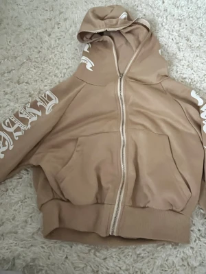 Beige hoodie med tryck och dragkedja - Snygg beige hoodie med vit tryckt text längs ärmarna och huvan. Hoodien har dragkedja framtill, två stora fickor och ribbade muddar. Perfekt för en avslappnad streetwear-look.
