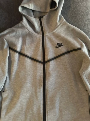 äkta Grå Nike zip hoodie   - Snygg grå zip hoodie från Nike med svarta detaljer och logga på bröstet. Tillverkad i mjukt material som känns skönt mot huden. Klassisk huva och dragkedja framtill gör den enkel att ta på och av. Perfekt för en sportig och avslappnad stil.  äkta Nike techfleece
