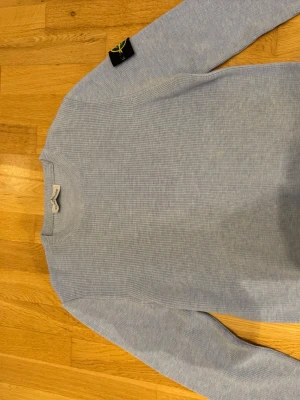 Ljusblå stickad tröja  - Snygg ljusblå stickad tröja från Stone Island med klassisk rund hals och ribbade muddar. Tröjan har den ikoniska Stone Island-loggan på vänster ärm. Perfekt för dig som gillar stilrena och trendiga plagg.