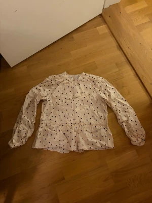 Prickig blus från Holly & Whyte XS - Söt ljusbeige blus med svarta prickar från Holly & Whyte by Lindex. Blusen har långärmade puffärmar, rund halsringning och volang nertill. Tunt och lätt material som ger en luftig känsla.