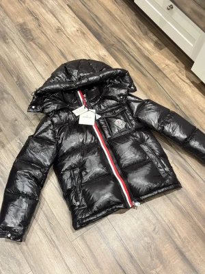 Svart Moncler pufferjacka med huva -  (Ljuset gör att den glänser mycket) Säljer en svart pufferjacka från Moncler med blank finish, huva och ikonisk logga på bröstet. Jackan har rödvit rand längs dragkedjan, praktiska fickor och innerfoder med serietidningsmotiv. Perfekt för kalla dagar och riktigt snygg streetstil.