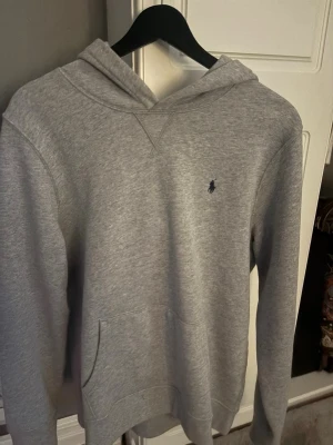 Grå hoodie från Polo Ralph Lauren xl i barn = S vuxen - Snygg grå hoodie från Polo Ralph Lauren med klassisk broderad logga på bröstet. Hoodien har huva, känguruficka och är tillverkad i mjukt bomullsmaterial. Perfekt för en avslappnad och stilren look.