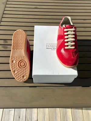 Maison Margiela röda sneakers - Maison Margiela sneakers i rött läder och mocka med beige snörning och gummisula. Klassisk low-top siluett med kontrasterande material och detaljer. Perfekta för dig som vill sticka ut med stilrena och trendiga skor.