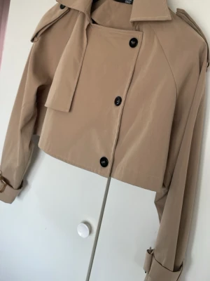 Kort beige trenchcoat från Dazy - Trendig kort trenchcoat i beige från Dazy med dubbelknäppning, axelklaffar och bälte vid ärmsluten. Jackan har klassisk krage och är tillverkad i ett lätt tyg som ger en snygg och stilren look.