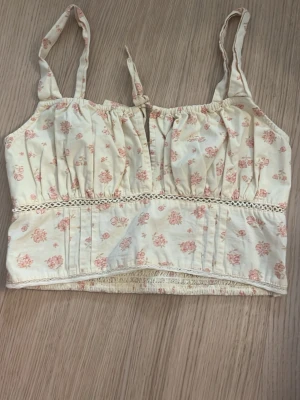 Blommig linnetopp med spetsdetalj - Söt ljusbeige linnetopp med rosa blommönster och smala axelband. Toppen har knyt framtill, spetsdetalj under bysten och smockad rygg för skön passform. Perfekt för varma dagar och en romantisk look.