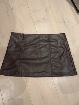 Brun skinnimitation kjol från H&M Divided - Snygg brun kortkjol i skinnimitation från H&M Divided. Kjolen har en rak passform och markerade sömmar framtill. Perfekt att styla med en stickad tröja eller t-shirt för en trendig look.