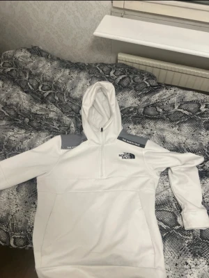 Vit hoodie från The North Face - Snygg vit hoodie från The North Face med grå detaljer på axlarna och logga på bröstet. Hoodien har huva och halv dragkedja framtill. Perfekt för dig som gillar sportig och clean stil. Materialet känns mjukt och bekvämt.