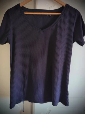 Svart ribbad t-shirt med v-ringning - Snygg svart t-shirt från The Black Label Collection i storlek M. T-shirten har ribbad struktur, korta ärmar och en v-ringad hals. Perfekt för en stilren och enkel look. Stor i storleken skön och sval.såg inte det ovanför tvättrådet och material förrän nu ❤️