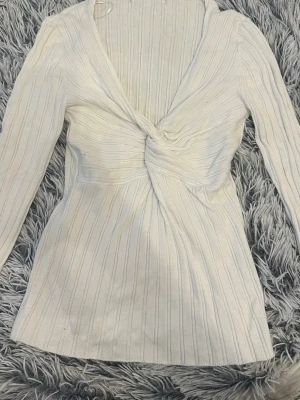 Vit ribbad knyttopp med lång ärm - Supersnygg vit ribbad topp med omlottdesign och lång ärm. Djup v-ringning och twistad detalj framtill ger en trendig look. Perfekt att styla med jeans eller kjol för en fräsch outfit.