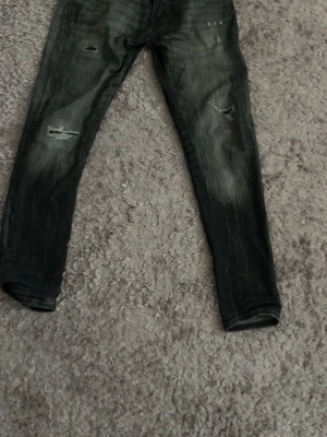 Svarta slitna jeans från Jack & Jones - Säljer ett par svarta jeans från Jack & Jones med slitningar på båda benen. Jeansen har en smal passform och klassisk femficksdesign. Materialet är denim med en tvättad look och detaljerade slitningar för en trendig stil.