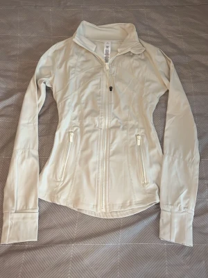 Vit träningsjacka från Lululemon - Snygg vit träningsjacka från Lululemon med figurnära passform, hög krage och dragkedja framtill. Jackan har två fickor med dragkedja och tumhål vid ärmsluten. Perfekt för löpning eller gym. Tillverkad i stretchigt funktionsmaterial.