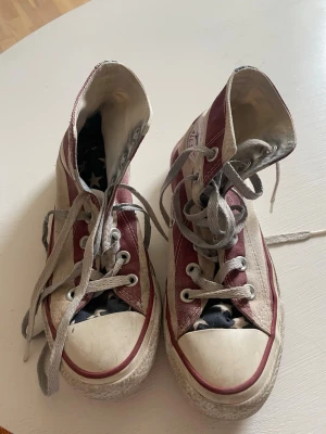 Converse All Star USA high tops - Säljer ett par Converse All Star high tops med amerikanskt flaggmönster i rött, vitt och blått. Skorna har vita gummisulor, grå snören och klassisk rund tå. Tillverkade i canvas med Converse-logga på sidan. Perfekta för dig som vill sticka ut med en unik sneaker.