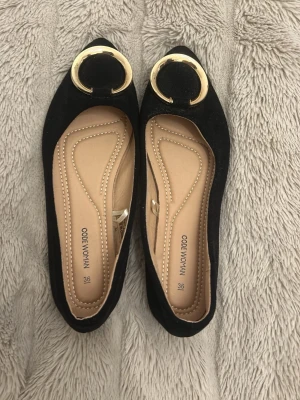 Svarta ballerinaskor med gulddetalj - Stilrena svarta ballerinaskor från Code Woman med spetsig tå och stor rund guldfärgad dekoration framtill. Skorna har en mjuk innersula i beige och ovandel i mockaimitation. Perfekta för dig som gillar en trendig och elegant look. Helt nya!