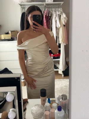 Champagnefärgad balklänning i satin från Meshki - Elegant långklänning i champagnefärgad satin med draperad omlottdetalj och oneshoulder-design. Klänningen har en mjuk, glansig yta och figurnära passform som ger en lyxig känsla. Perfekt för speciella tillfällen. Helt ny balklänning från meshki, den heter ”Daila Off Shoulder Satin Maxi Dress - Blush Gold ” i storlek XS men ska varna att den är liten i storleken men detta är runt om midjan och bysten eftersom det är en korsett. Men en väldig fin balklänning och längden behöver man inte vara oroli