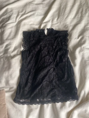 Svart spetsblus med hög hals - Snygg svart blus i spets med hög hals och ärmlös design. Blusen har fina spetsdetaljer längs kanterna och en liten öppning med knapp i nacken. Perfekt för dig som gillar en elegant och trendig look.