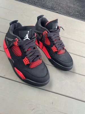 Svarta och röda Air Jordan 4 sneakers - Säljer ett par Air Jordan 4 Red Thunders, sneakers i svart och rött med klassisk Jumpman-logga på plösen och sulan. Skorna har svarta skosnören, röd mesh och detaljer samt en robust sula med rött och svart mönster. Perfekta för dig som gillar streetwear och sportig stil.
