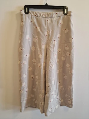 Beiga blommiga byxor från Zara - Beiga vida trekvartsbyxor från Zara Basic med vitt och rött blommönster. Byxorna har lös passform, hög midja och är gjorda i ett lätt tyg som ger en chill och trendig känsla. Perfekt för dig som gillar unika detaljer och vill sticka ut lite extra.