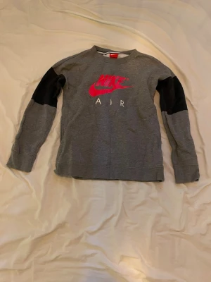 Grå Nike Air sweatshirt med svarta detaljer - Snygg grå sweatshirt från Nike med stort rött Nike Air-tryck på bröstet och svarta paneler på ärmarna. Tröjan har rund halsringning och är tillverkad i mjukt bomullsmaterial. Perfekt för en sportig och avslappnad stil.