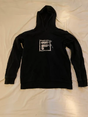 Svart hoodie med FILA-logga - Svart hoodie från FILA med stor vit logga i handritad stil på bröstet. Klassisk passform med huva och långa ärmar. Tillverkad i mjukt bomullsmaterial som känns skönt mot huden. Perfekt för dig som gillar streetwear och vill ha en enkel men snygg look.