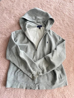 Grå hoodie från Polo Ralph Lauren - Snygg grå hoodie från Polo Ralph Lauren med dragkedja framtill och klassisk blå logga broderad på bröstet. Hoodien har huva och två fickor fram. Tillverkad i mjuk bomullsblandning som känns skön mot huden. Perfekt för en avslappnad och stilren look.Obs litet knappt synbart hål vid bröstet och ett vid vänstra handleden. Tröjan är XS men passar också S