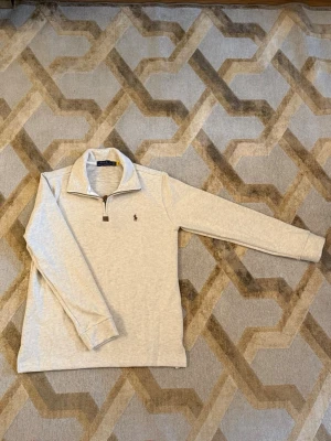 Ljusgrå stickad tröja från Polo Ralph Lauren - Ljusgrå stickad tröja från Polo Ralph Lauren med halv dragkedja och klassisk krage. Tröjan har ett brunt broderat logomärke på bröstet och ribbade muddar vid ärmslut. Perfekt för lager-på-lager och snygg till jeans.