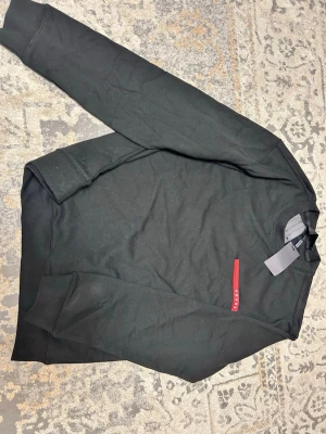 Svart sweatshirt från Prada - Cool svart sweatshirt från Prada med klassisk rund halsringning och lång ärm. På bröstet finns en röd detalj med Prada-loggan. Tröjan är tillverkad i mjukt material och har ribbade muddar vid ärmslut och nederkant. Perfekt för en stilren look.