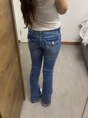 Blå bootcut jeans med fickdetaljer - Snygga blå bootcut jeans med vita knappar på bakfickorna och slitningar framtill. Jeansen har låg midja och klassisk femficksdesign. Perfekta för dig som gillar en avslappnad och trendig look.