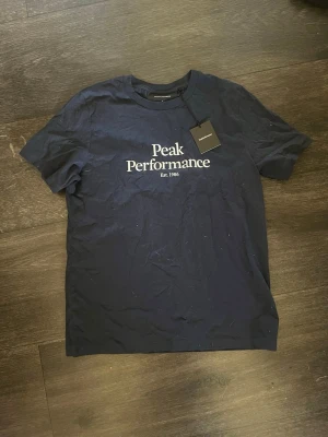 Mörkblå Peak Performance t-shirt - Snygg mörkblå t-shirt från Peak Performance med vit logga tryckt på bröstet. Klassisk rund hals och korta ärmar. Perfekt för en stilren och avslappnad look. Tillverkad i mjuk bomull för maximal komfort.   Kan diskutera priser