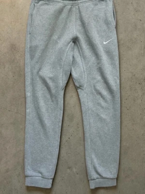 Gråa mjukisbyxor från Nike - Snygga grå mjukisbyxor från Nike med klassisk vit logga på vänster lår. Byxorna har resår i midjan, muddar vid bensluten och sidofickor samt en bakficka. Tillverkade i mjukt sweatshirtmaterial som är perfekt för chill eller träning. Tröja finns !!!