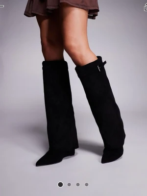 Svarta höga mockastövlar med spänne - Snygga svarta stövlar i mocka med högt skaft och dekorativt spänne upptill. Stövlarna har spetsig tå och låg klack, vilket ger en trendig och stilren look. Perfekta att bära till kjol eller klänning för en cool vibe. HELT OANVÄNDA