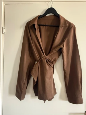 Brun omlottblus med knytband - Snygg brun omlottblus med långa ärmar och bred krage. Blusen har ett knytband i midjan som ger en smickrande siluett och är tillverkad i ett glansigt, mjukt material som känns lyxigt. Perfekt att styla med jeans eller kjol.