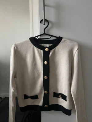 Beige och svart kofta med guldknappar - Stilren kofta i beige med svarta detaljer och dekorativa guldknappar framtill. Rund halsringning, långa ärmar. Perfekt för en chic och klassisk look. Storlek S