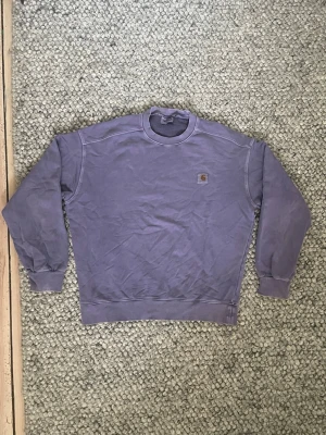 Lila sweatshirt från Carhartt - Lila sweatshirt från Carhartt med rund halsringning och liten logopatch på bröstet. Strl L
