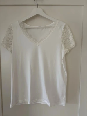 Vit topp med spetsdetaljer - Snygg vit topp med v-ringning och spetsdetaljer runt halsen samt spetsärmar. Kortärmad modell i mjukt material som känns både stilren och feminin. Perfekt att matcha med jeans eller kjol för en fräsch look. Lös passform som faller snyggt. Axelbredd 36 cm och längd 62 cm. Lapparna bortklippta.
