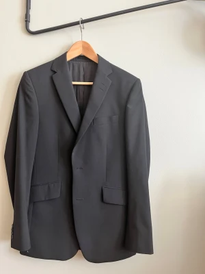 Svart kavaj från Bläck, MQ - Stilren svart kavaj från Bläck med klassisk passform och två framfickor med lock. Perfekt för en dressad look och funkar till skjorta eller t-shirt. Regular fit. 