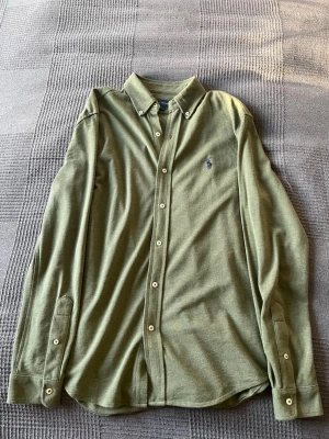 Olivgrön skjorta från Polo Ralph Lauren - Snygg olivgrön skjorta från Polo Ralph Lauren med klassisk krage och knappar framtill. Skjortan har lång ärm och den ikoniska loggan broderad på bröstet. Tillverkad i mjuk bomull som känns skön mot huden. Perfekt för en clean och stilren look.