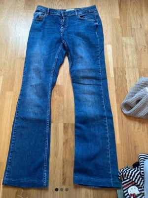 Blå bootcut jeans med klassisk passform - Säljer ett par blå bootcut jeans med klassisk femficksdesign och normalhög midja. Jeansen har en snygg tvätt och lätt utsvängda ben, perfekta för en avslappnad och trendig look. Materialet är mjukt denim som ger en skön känsla hela dagen. Jag tror storleken är M/L