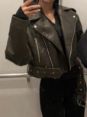 Grön cropped skinnjacka Zara - Grön cropped skinnjacka från Zara med breda slag, silvriga dragkedjor och bälte i midjan. Jackan har två framfickor med dragkedja och axelklaffar. 