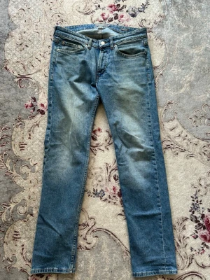 Blå raka jeans med klassisk tvätt - Säljer ett par blå jeans med rak passform och klassisk femficksdesign. Jeansen har en snygg ljusblå tvätt med subtila slitningar framtill och bakfickor. Perfekta för en avslappnad och stilren look.