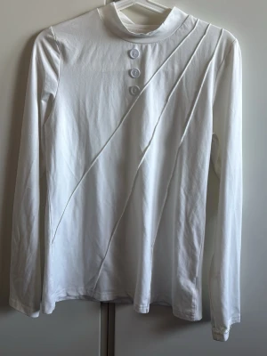 Vit långärmad polotopp med knappar - Vit långärmad polotopp med tre dekorativa knappar framtill och diskreta diagonala sömmar som ger en cool look. Toppens material känns mjukt och stretchigt, perfekt för att matcha med jeans eller kjol. Enkel och stilren design som passar till många outfits.