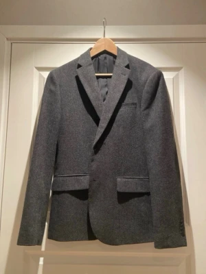 Mörkgrå ullkavaj med armbågslappar - Stilren mörkgrå kavaj i ullmix med klassisk passform och svarta armbågslappar. Två framfickor med lock, bröstficka och knappar vid ärmslut. Perfekt för en dressad look och funkar året runt.