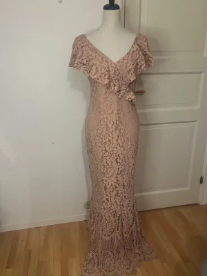 Lång spetsklänning med volang - Romantisk långklänning i ljusrosa spets med v-ringning och bred volang över axlarna. Klänningen har figurnära passform och ett vackert blommigt spetsmönster. Perfekt för speciella tillfällen när du vill känna dig extra fin.