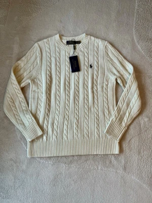Vit kabelstickad tröja  - Vit klassisk kabelstickad tröja. Med rund halsring och en mörkblå broderad logga på bröstet. Helt ny tröja, aldrig använd. Säljer pga att den är för stor för mig. 
