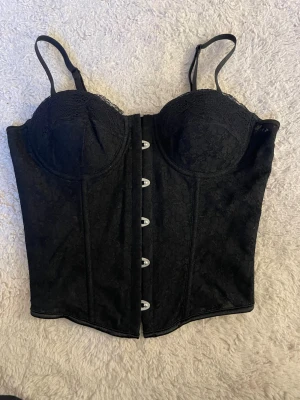 Svart spetskorsett med snörning - Elegant svart korsett med spetsdetaljer och formade kupor. Framsidan har metallknappar och baksidan har snörning med breda satinband. Justerbara axelband och diskret blommönster i tyget💗