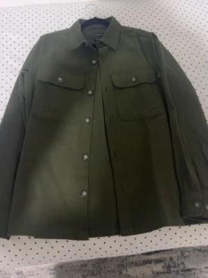 Olivgrön overshirt från S.C.W. - Stilren olivgrön overshirt från S.C.W. i återvunnet material. Skjortjackan har två stora bröstfickor med lock och knappar, klassisk krage och knäppning framtill. Perfekt lager på lager-plagg för en trendig look.