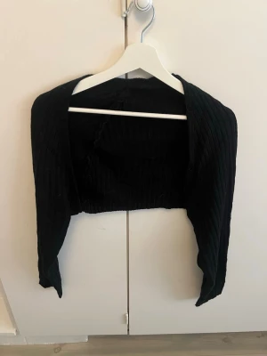 Svart ribbad bolero i XS - Säljer en svart ribbad bolero i storlek XS. Kort modell med långa ärmar och öppen front, perfekt att slänga över en topp eller klänning. Materialet är mjukt och stretchigt för en bekväm passform.