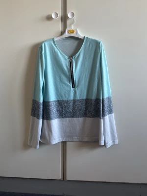 Ljusblå långärmad topp med dragkedja - Långärmad topp i ljusblått med breda grå och vita block längst ner. Toppen har en rund halsringning och en snygg dragkedja framtill. Perfekt för en chill och avslappnad stil.